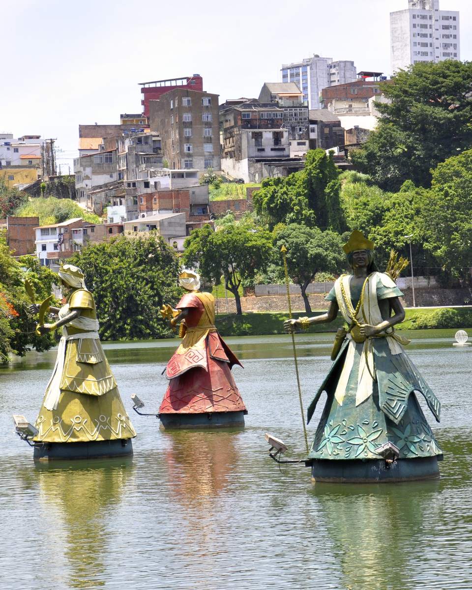 Esculturas dos orixás sobre a água do Dique do Tororó, em Salvador, com árvores e prédios ao fundo.