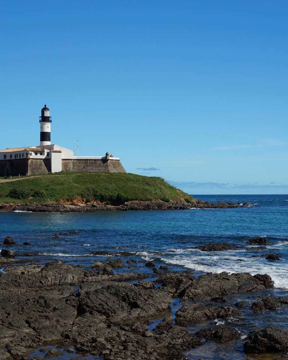 Farol da Barra e o Forte de Santo Antônio sobre o morro verde, em Salvador, vistos das pedras escuras da praia, com ondas brancas quebrando no mar azul e o céu limpo.