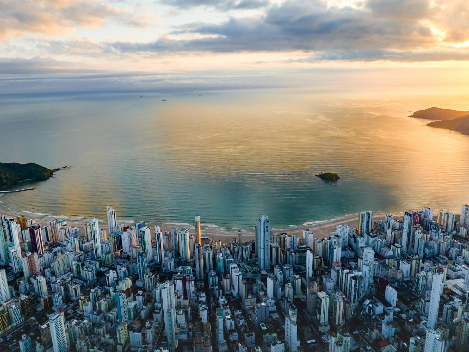 Balneário Camboriú vista do alto ao nascer do sol, com os prédios altos da orla em primeiro plano, a baía calma refletindo o céu dourado e a ilha no meio do mar.