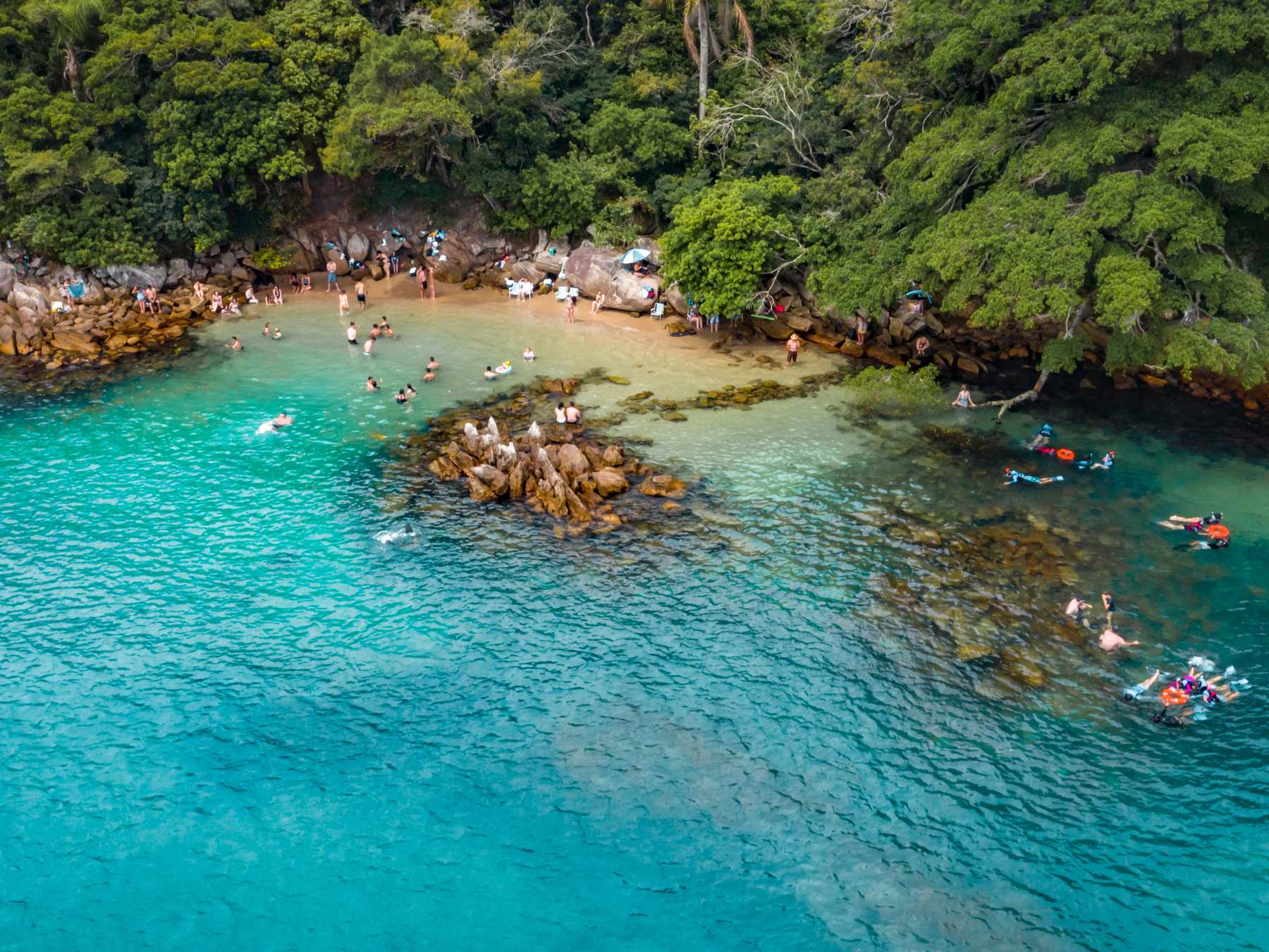 Enseada de água azul-turquesa e transparente em Bombinhas vista do alto, com caiaques e banhistas entre as pedras e a mata atlântica descendo até o mar.