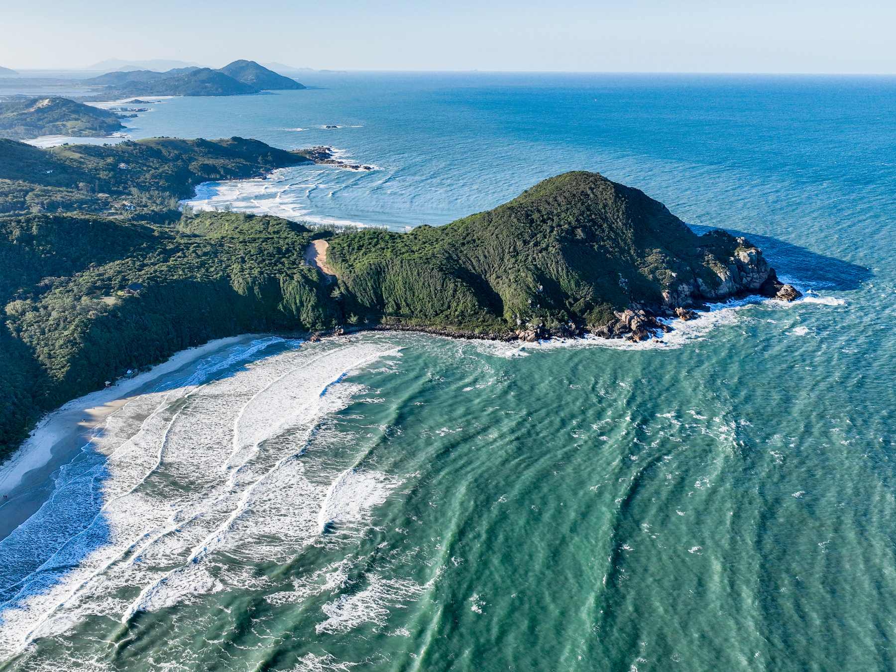 Praia do Rosa vista do alto, com ondas brancas quebrando na areia, o morro verde entrando no mar azul e a costa recortada ao fundo.