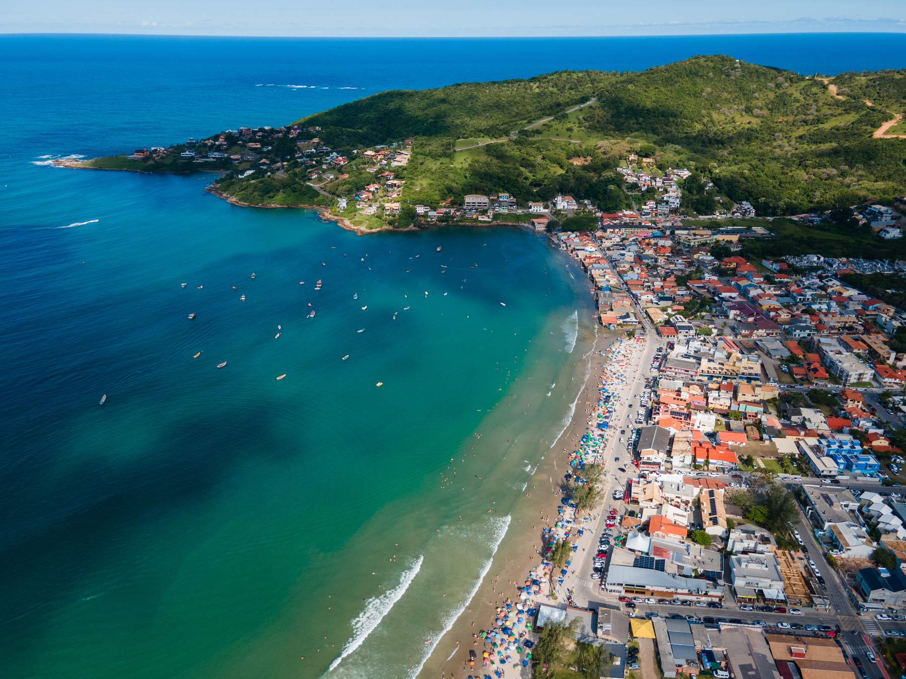 Vista aérea da enseada de Garopaba, com o mar azul, barcos ancorados, a faixa de areia e a cidade de casas coloridas ao pé do morro verde.