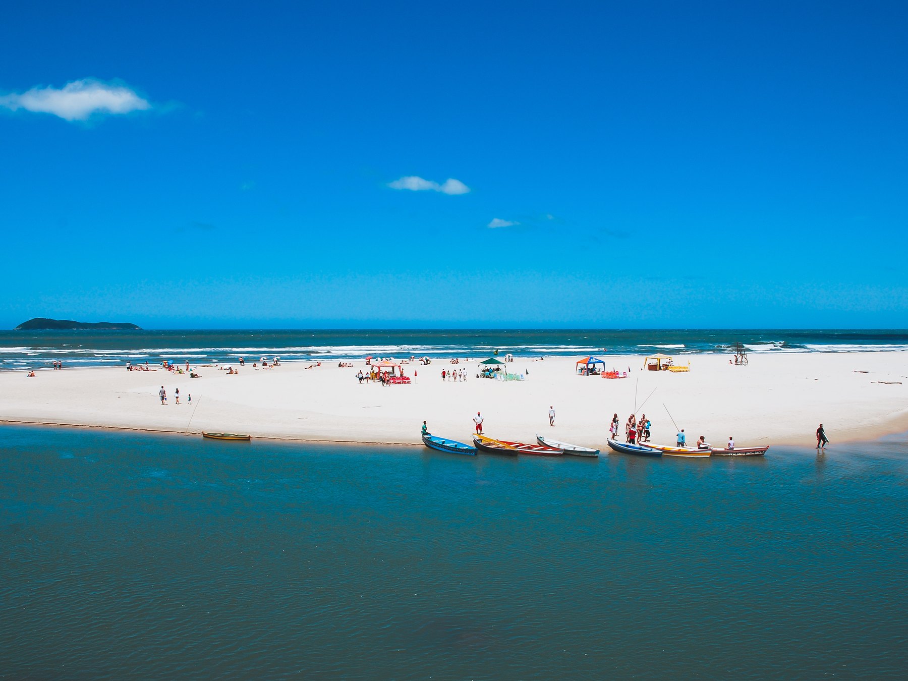 Barcos de pesca coloridos na foz do rio da Guarda do Embaú, com banhistas na faixa de areia branca e o mar azul ao fundo sob o céu limpo.