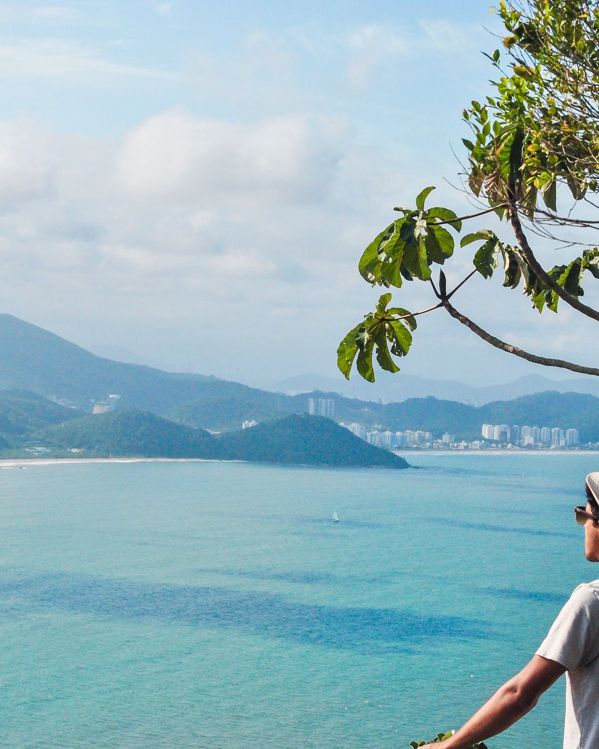 Rapaz de chapéu e camiseta branca olhando do alto de um morro para o mar verde-azulado e os prédios de Balneário Camboriú ao fundo.
