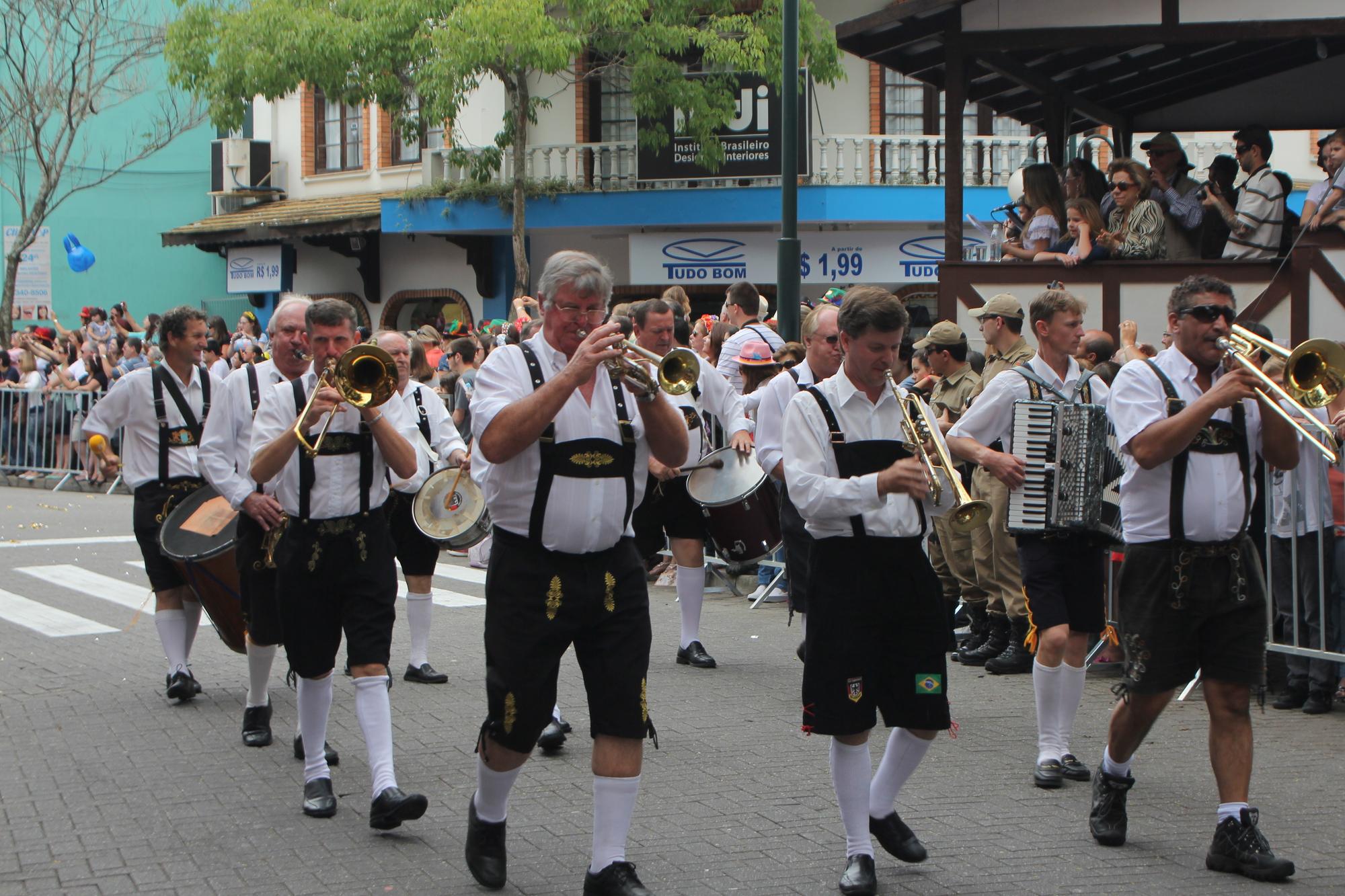 Banda de músicos de bermuda de couro, suspensórios e meias brancas tocando trompete, trombone, acordeão e tambor no desfile da Oktoberfest, em Blumenau, com o público atrás da grade e num camarote de madeira.