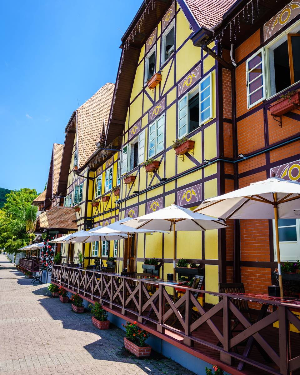 Fachada em enxaimel amarela e vermelha de um restaurante na Vila Germânica, em Blumenau, com guarda-sóis brancos, varanda de madeira e o céu azul.