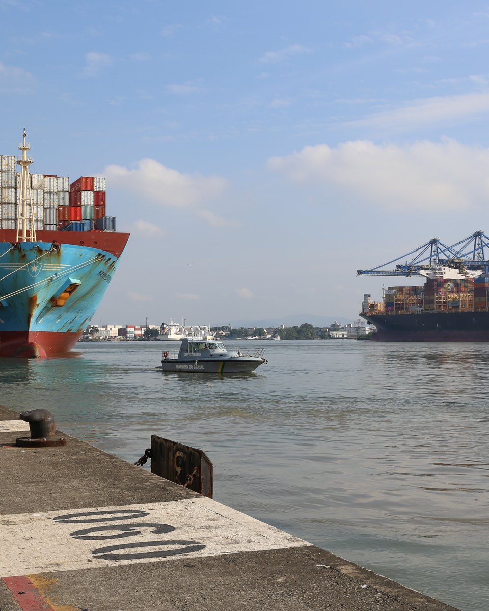 Navio porta-contêineres atracado no cais do Porto de Itajaí, com guindastes de carga ao fundo, outro navio no rio Itajaí-Açu e um barco de apoio na água.
