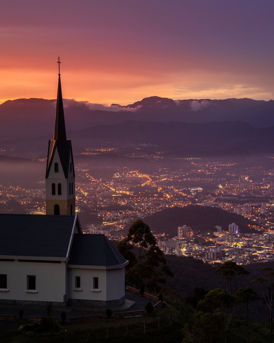 Capela alpina de torre pontiaguda no alto de um morro em Jaraguá do Sul, ao entardecer, com as luzes da cidade no vale e as montanhas contra o céu laranja e lilás.