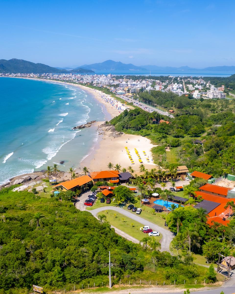 Vista aérea da Praia de Mariscal, em Bombinhas, com a faixa de areia curva, o mar verde-azulado, uma pousada de telhados vermelhos entre a mata e a estrada chegando à praia.