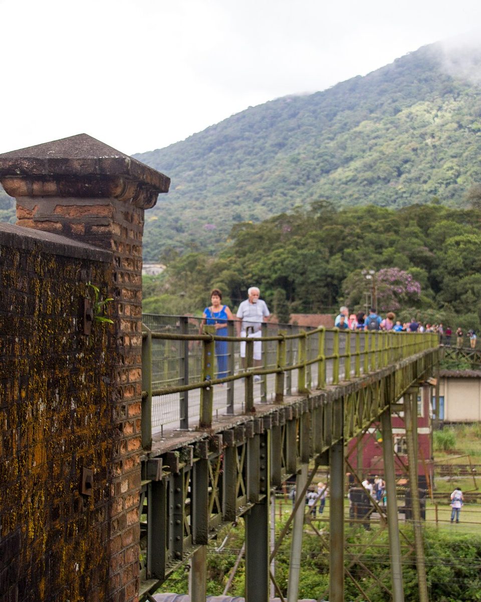 Visitantes na passarela de ferro sobre o pátio ferroviário de Paranapiacaba, com a serra coberta de mata ao fundo.