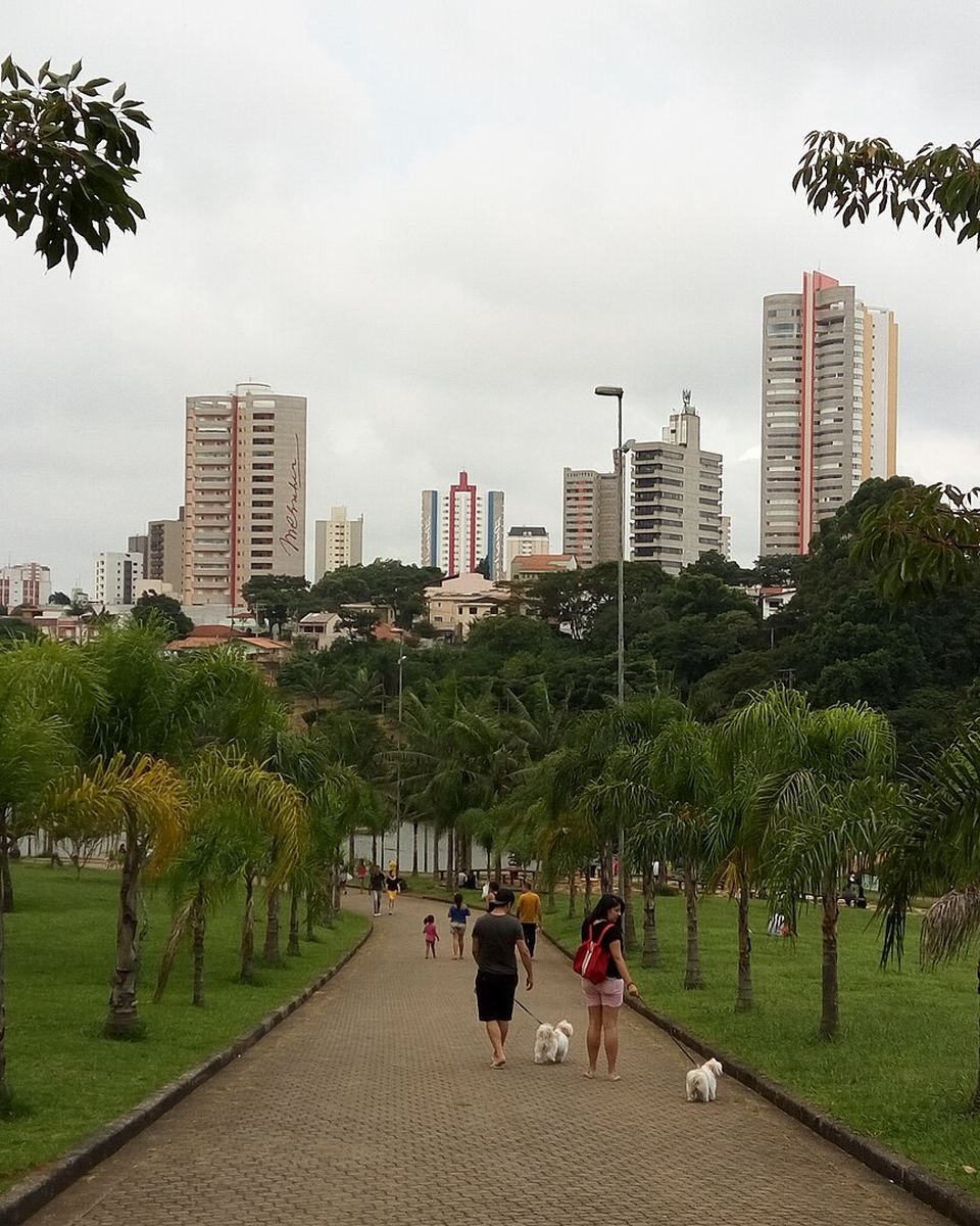 Pessoas caminhando por uma alameda do Parque Central de Santo André, com prédios ao fundo.