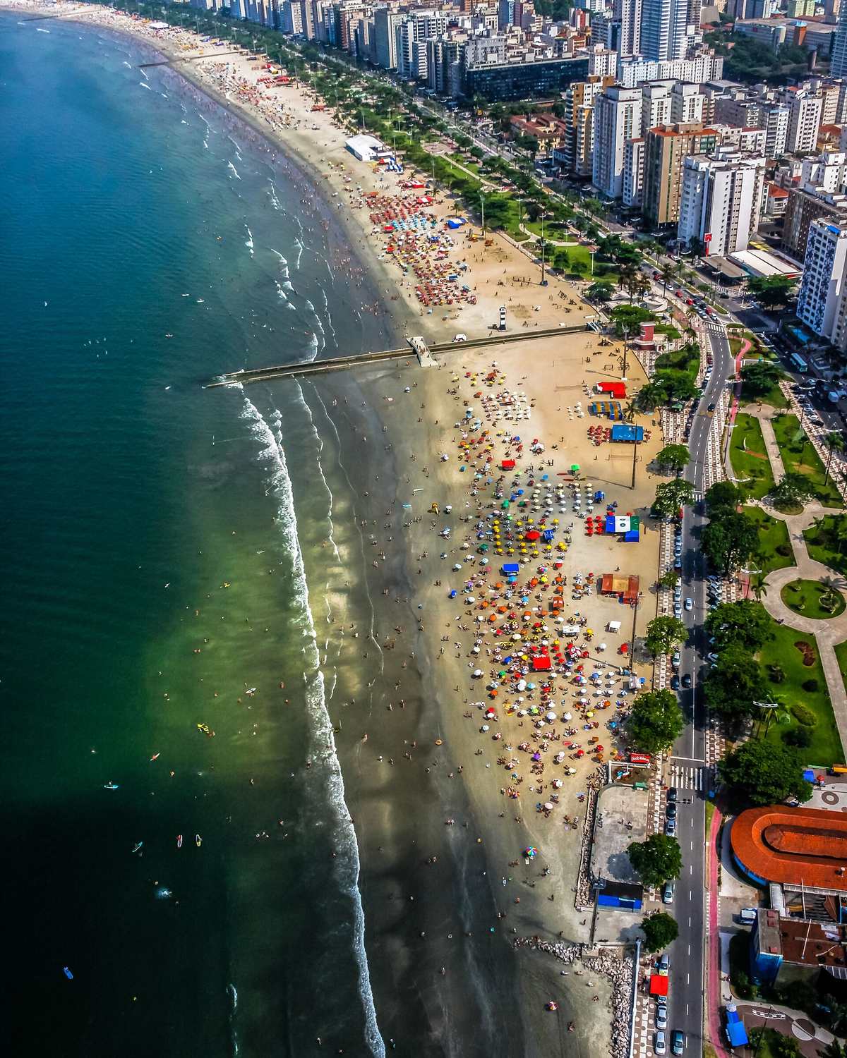 Vista aérea da praia de Santos cheia de guarda-sóis, com o jardim da orla e os prédios ao longo da avenida.