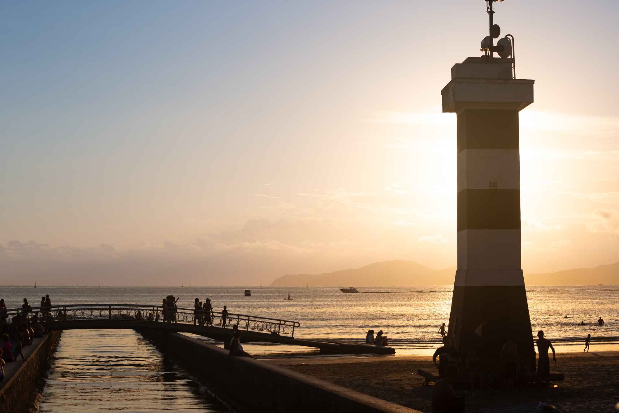 Pessoas caminhando na passarela e na areia ao lado do farol listrado do canal 6, em Santos, ao pôr do sol.