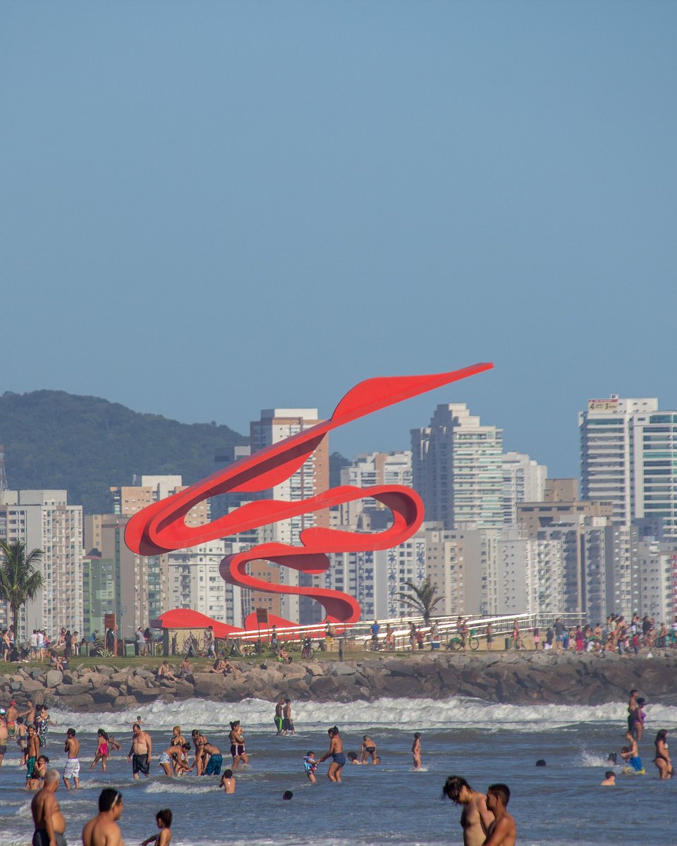 Banhistas no mar do José Menino, em Santos, com uma escultura vermelha e os prédios da orla ao fundo.