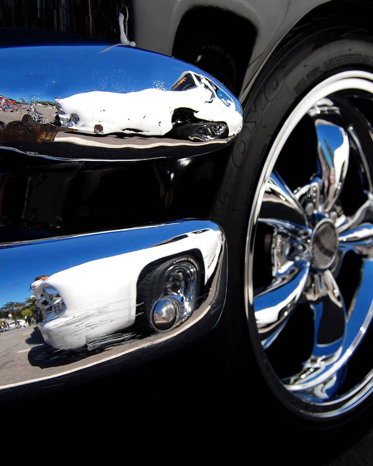 Fotógrafo agachado refletido no para-choque cromado de um carro antigo, ao lado da roda, numa exposição de carros em São Bernardo do Campo.