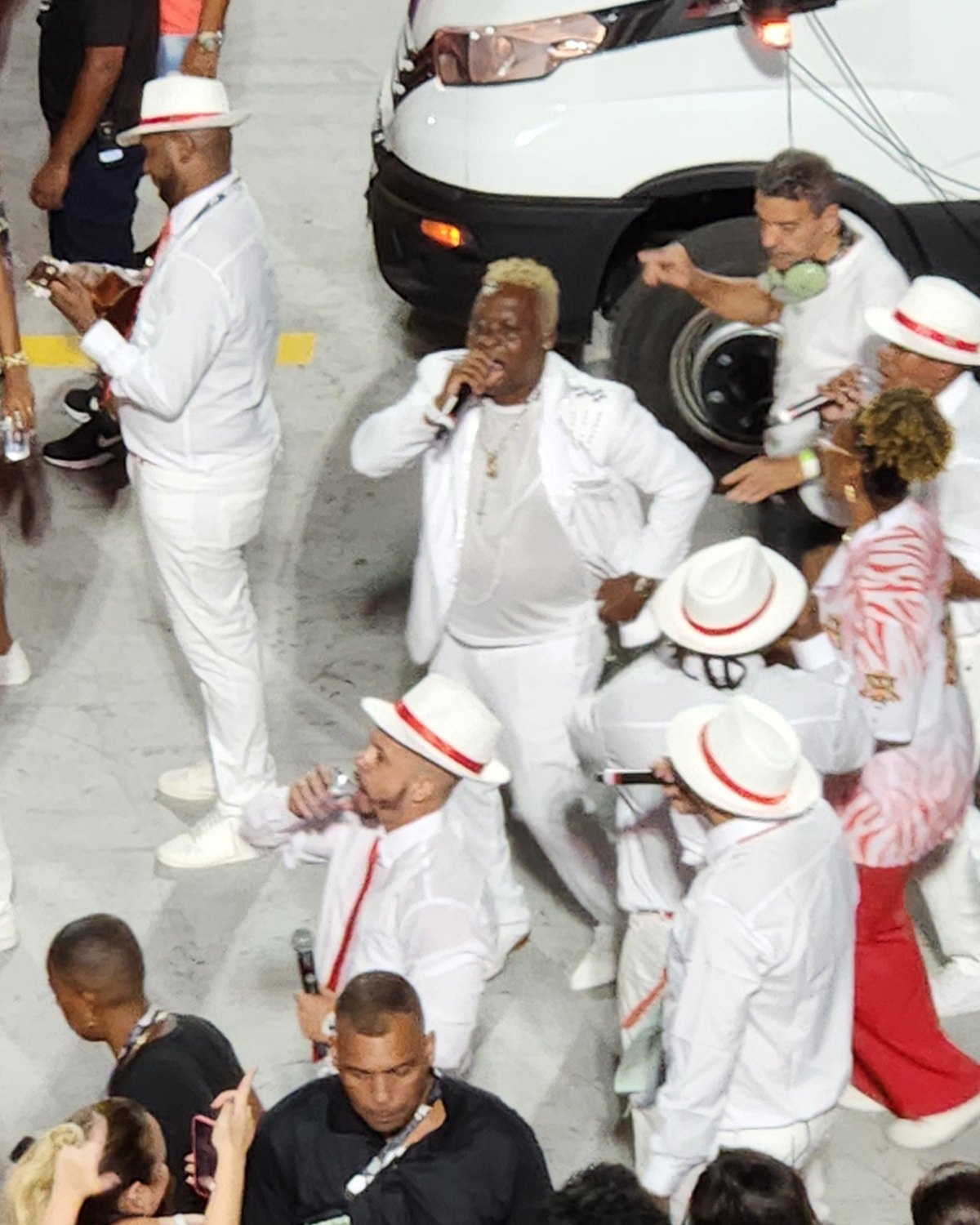 Sambistas de terno branco e chapéu panamá com fita vermelha cantando e tocando cavaquinho ao lado de ritmistas de roupa laranja e cocar amarelo, no desfile da Unidos do Porto da Pedra, escola de samba de São Gonçalo.