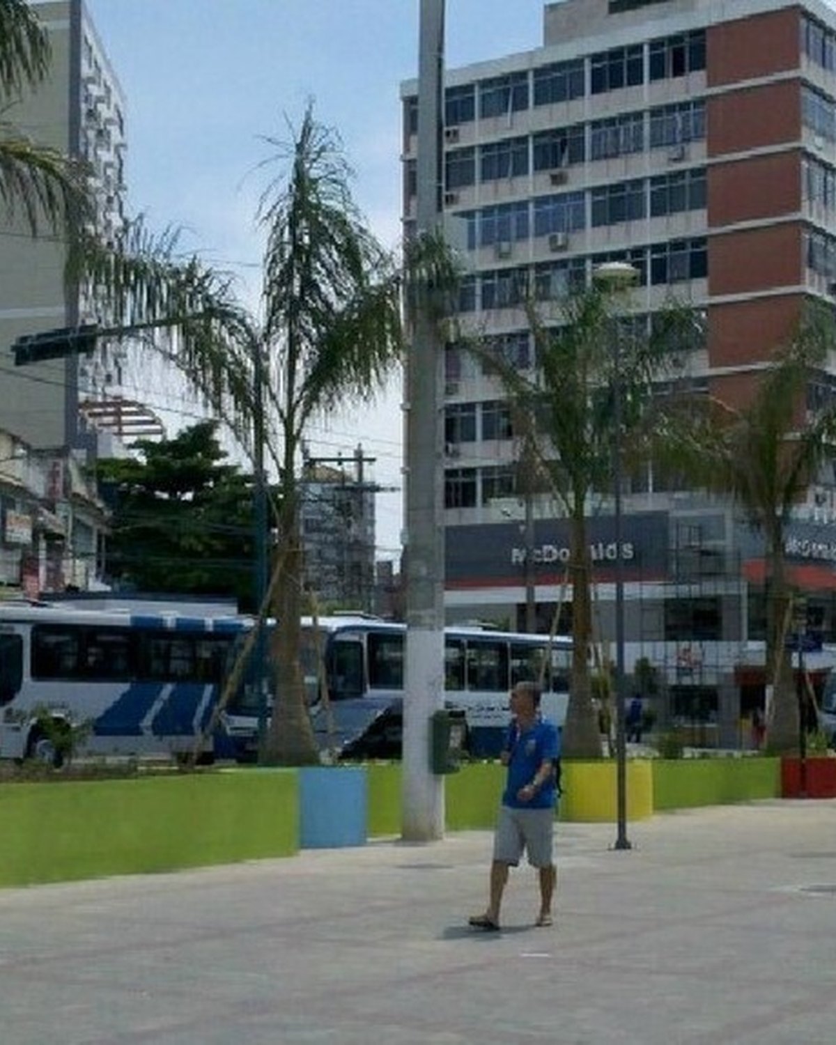 Praça no Centro de São Gonçalo num dia de sol, com canteiros coloridos, palmeiras, um homem de camiseta azul caminhando, pedestres na calçada, ônibus parados ao fundo e prédios comerciais.