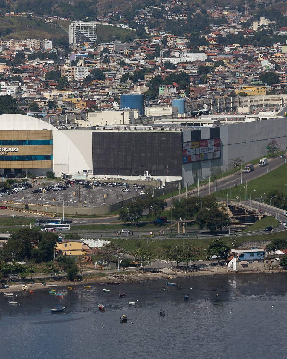 Vista aérea do São Gonçalo Shopping, prédio branco e cinza com estacionamento, cercado de bairros de casas e prédios baixos, com barcos de pesca na beira da Baía de Guanabara em primeiro plano.