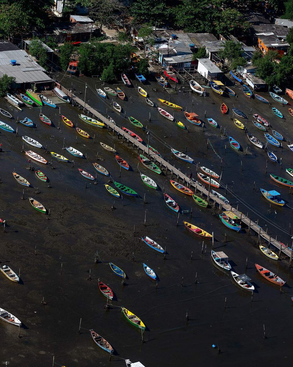 Vista aérea de dezenas de barcos de pesca coloridos parados na lama da maré baixa, na beira da Baía de Guanabara, em São Gonçalo, com um píer de madeira e casas entre as árvores.
