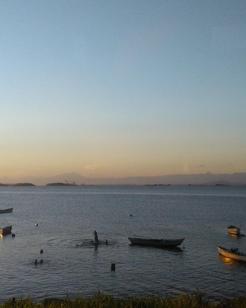 Baía de Guanabara ao entardecer, vista da margem de São Gonçalo, com pequenos barcos de pesca espalhados na água calma e montanhas no horizonte.
