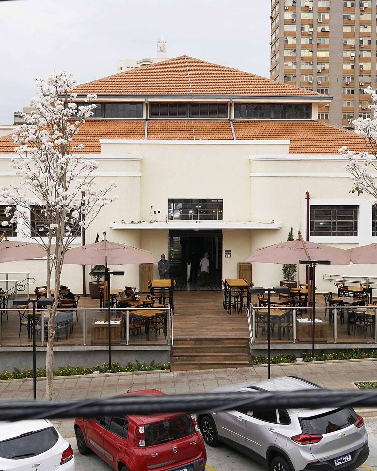 Fachada branca do Mercado Municipal de São José do Rio Preto, de 1944, com mesas e guarda-sóis na varanda, carros estacionados e prédios ao fundo.
