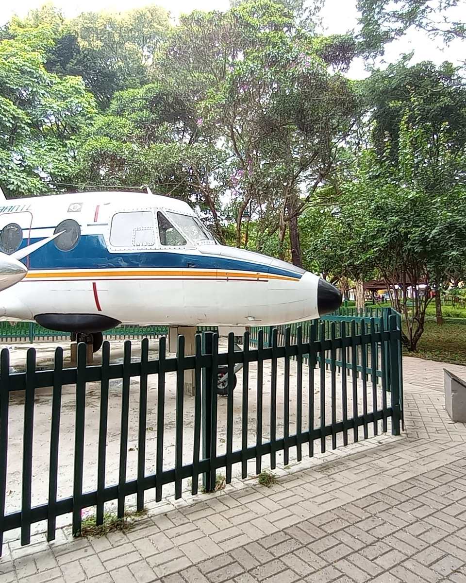 Pessoas caminhando ao lado de um avião bimotor branco e azul exposto no Parque Santos Dumont, em São José dos Campos, cercado por árvores.