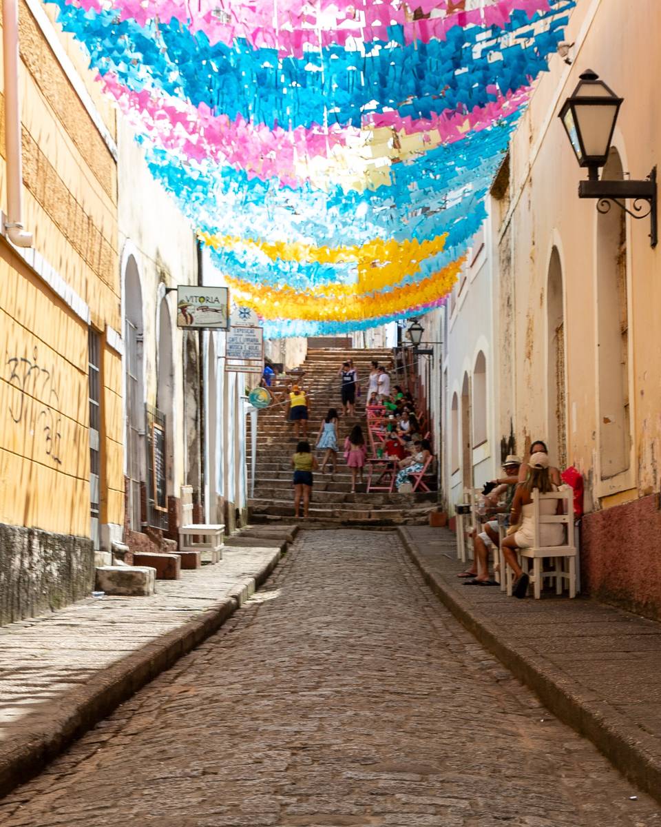 Rua de pedra do Beco Catarina Mina coberta por bandeirinhas de papel azuis, rosas e amarelas, com pessoas sentadas e subindo a escadaria ao fundo