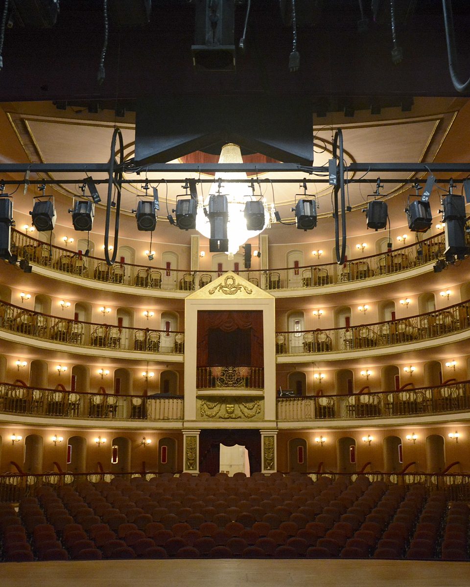 Interior do Teatro Arthur Azevedo visto do palco, com as galerias curvas de camarotes iluminadas, o lustre central e a plateia de poltronas vermelhas