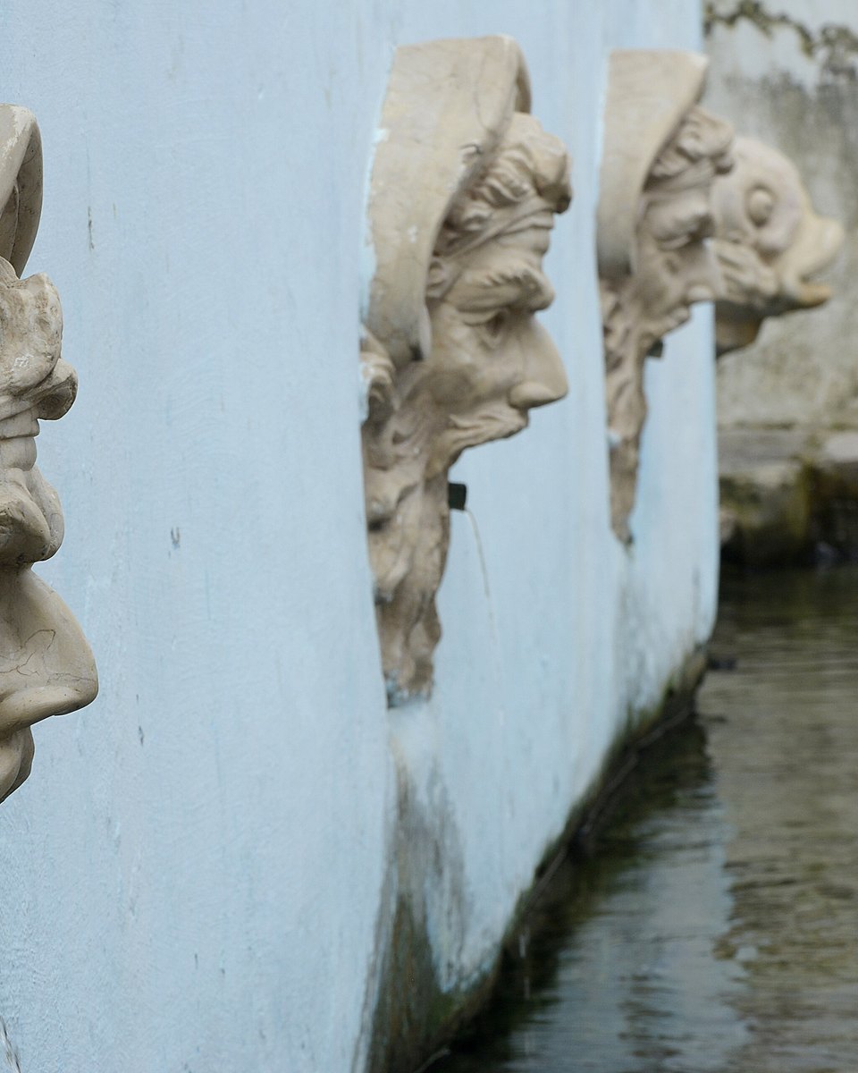 Mascarões de pedra esculpidos na parede branca da Fonte do Ribeirão, com a água correndo no canal logo abaixo