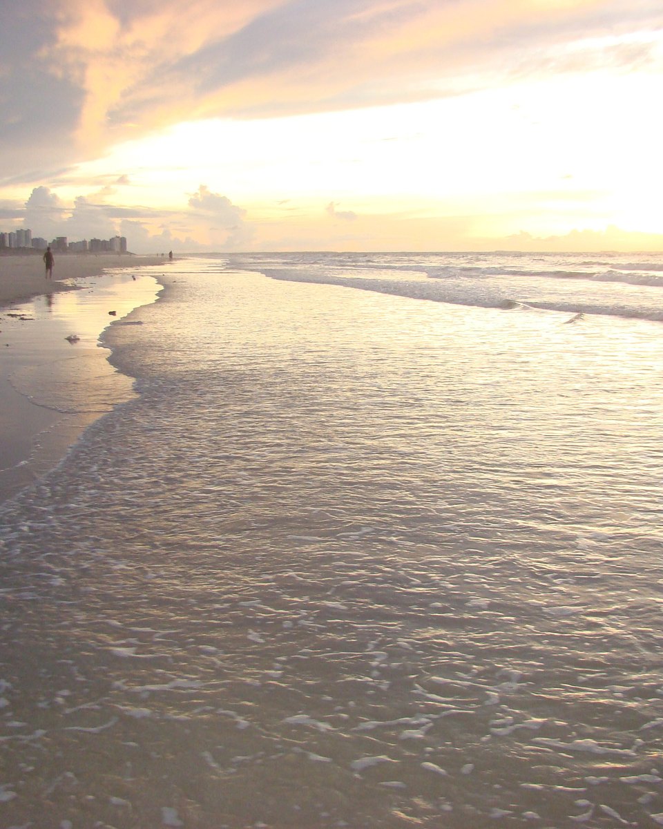 Pôr do sol na Praia do Calhau, com o sol baixo sobre o mar, a areia molhada refletindo a luz dourada e pessoas caminhando ao longe