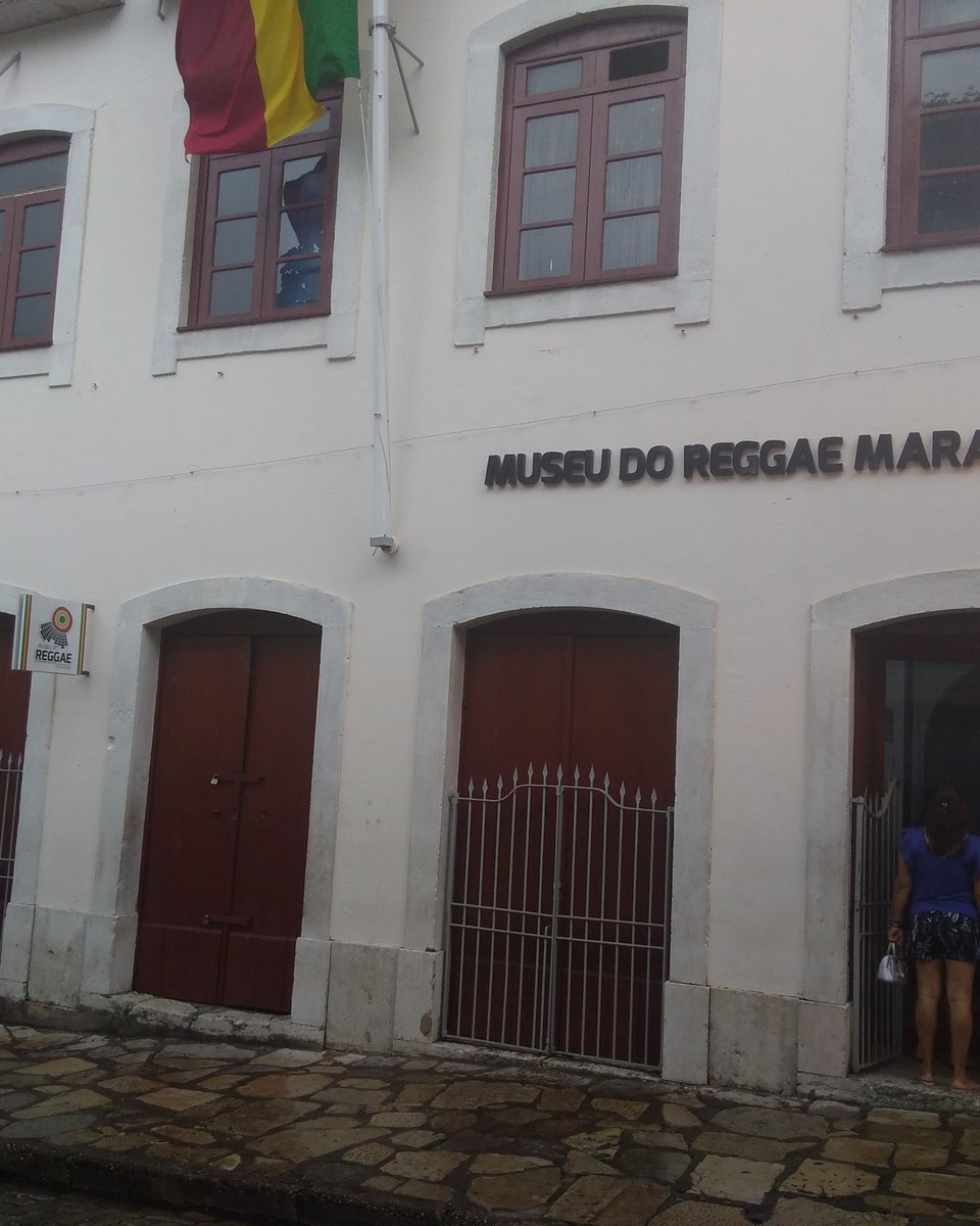 Fachada branca do Museu do Reggae do Maranhão, com portas vermelhas, letreiro na parede e pessoas na entrada