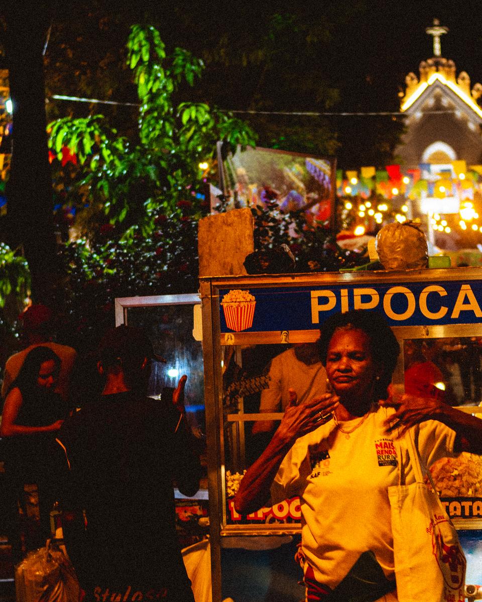 Barraca de pipoca iluminada em uma festa de São João em São Luís, com a vendedora atrás do carrinho, bandeirinhas coloridas no alto e gente circulando ao redor