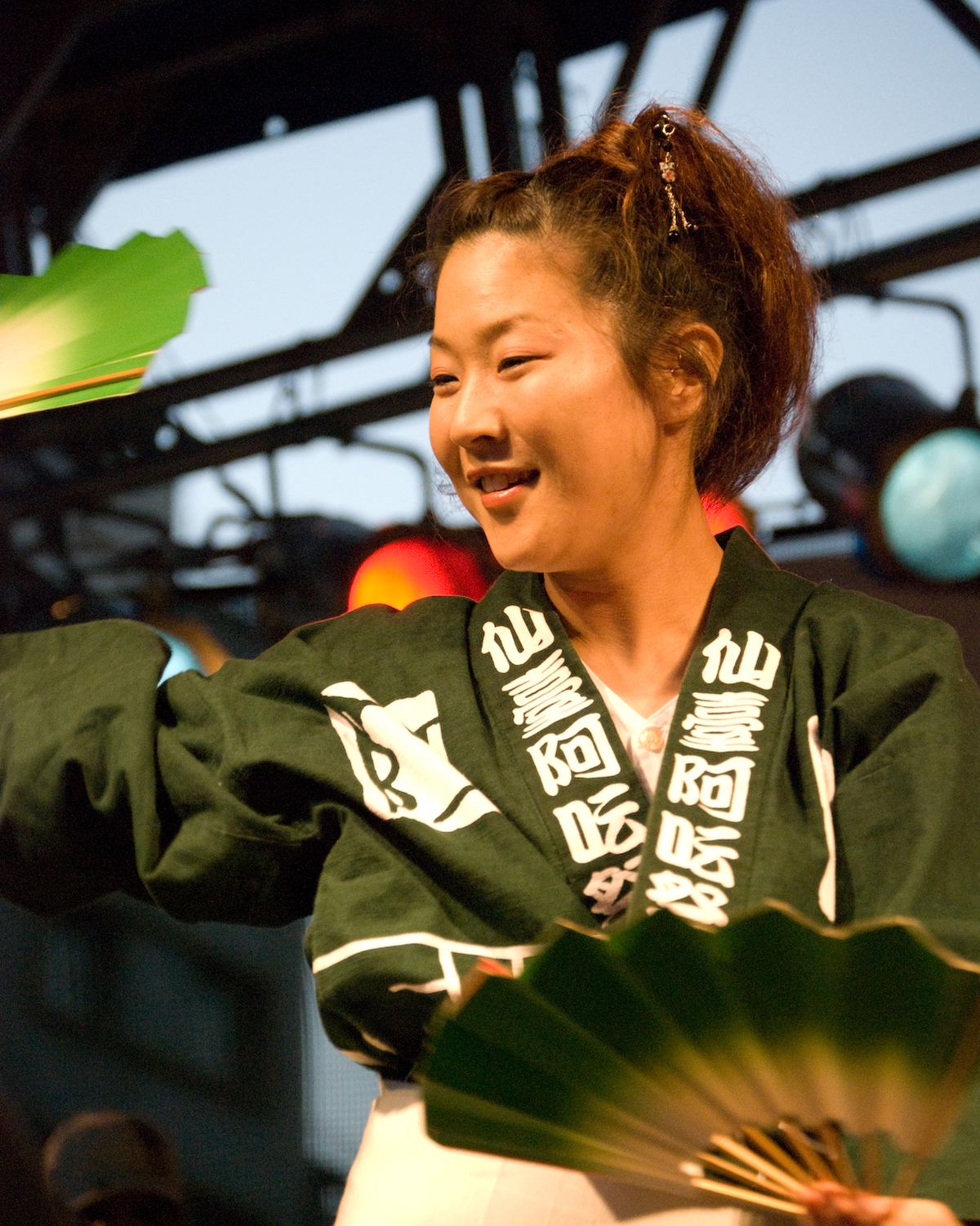 Dançarina de casaco tradicional japonês verde sorrindo e girando um leque durante o Tanabata Matsuri, na Liberdade.