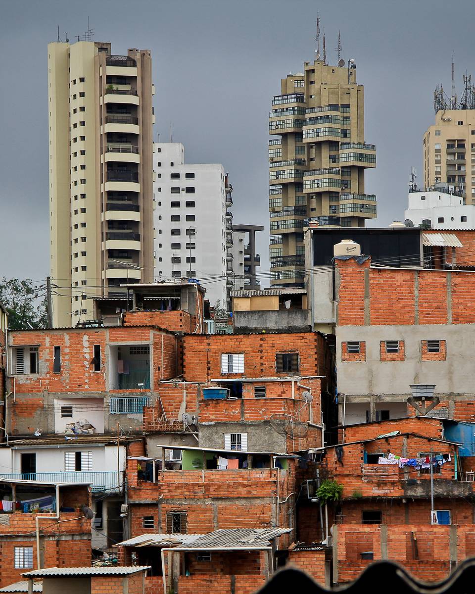 Casas de tijolo de Paraisópolis em primeiro plano e prédios altos ao fundo, sob céu nublado.