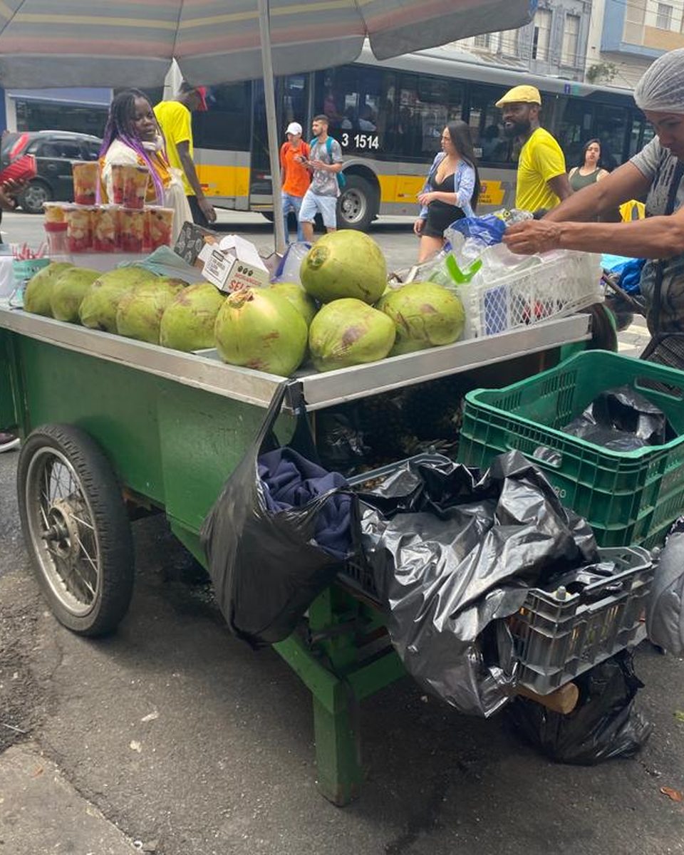 Vendedor ambulante arrumando cocos num carrinho numa rua movimentada do Brás, com pedestres e um ônibus ao fundo.