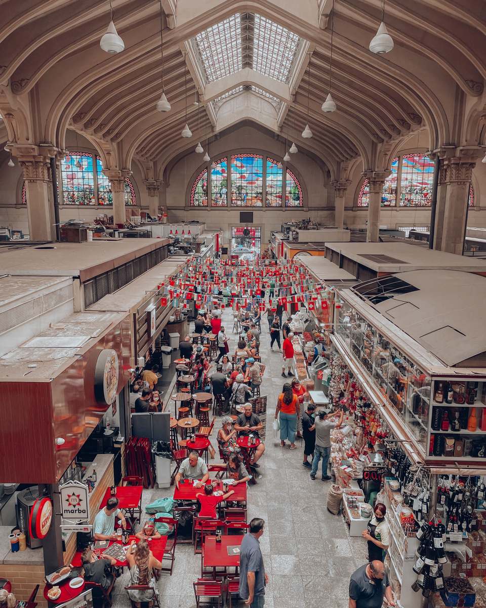 Vista do alto do corredor central do Mercado Municipal de São Paulo, cheio de gente entre bancas e mesas vermelhas, com os vitrais coloridos e o teto em arcos ao fundo.