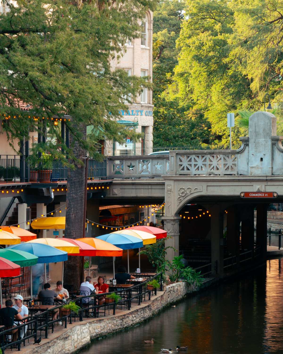 Gente comendo sob guarda-sóis coloridos à beira do River Walk, ao lado da ponte da Commerce Street, em San Antonio.