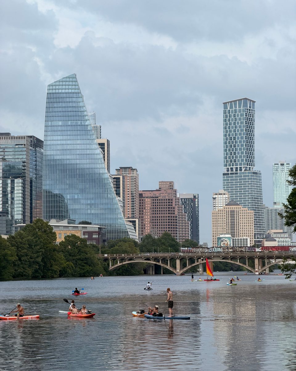 Pessoas de caiaque e stand up paddle no Lady Bird Lake, com a ponte da Congress Avenue e os prédios do centro de Austin ao fundo.