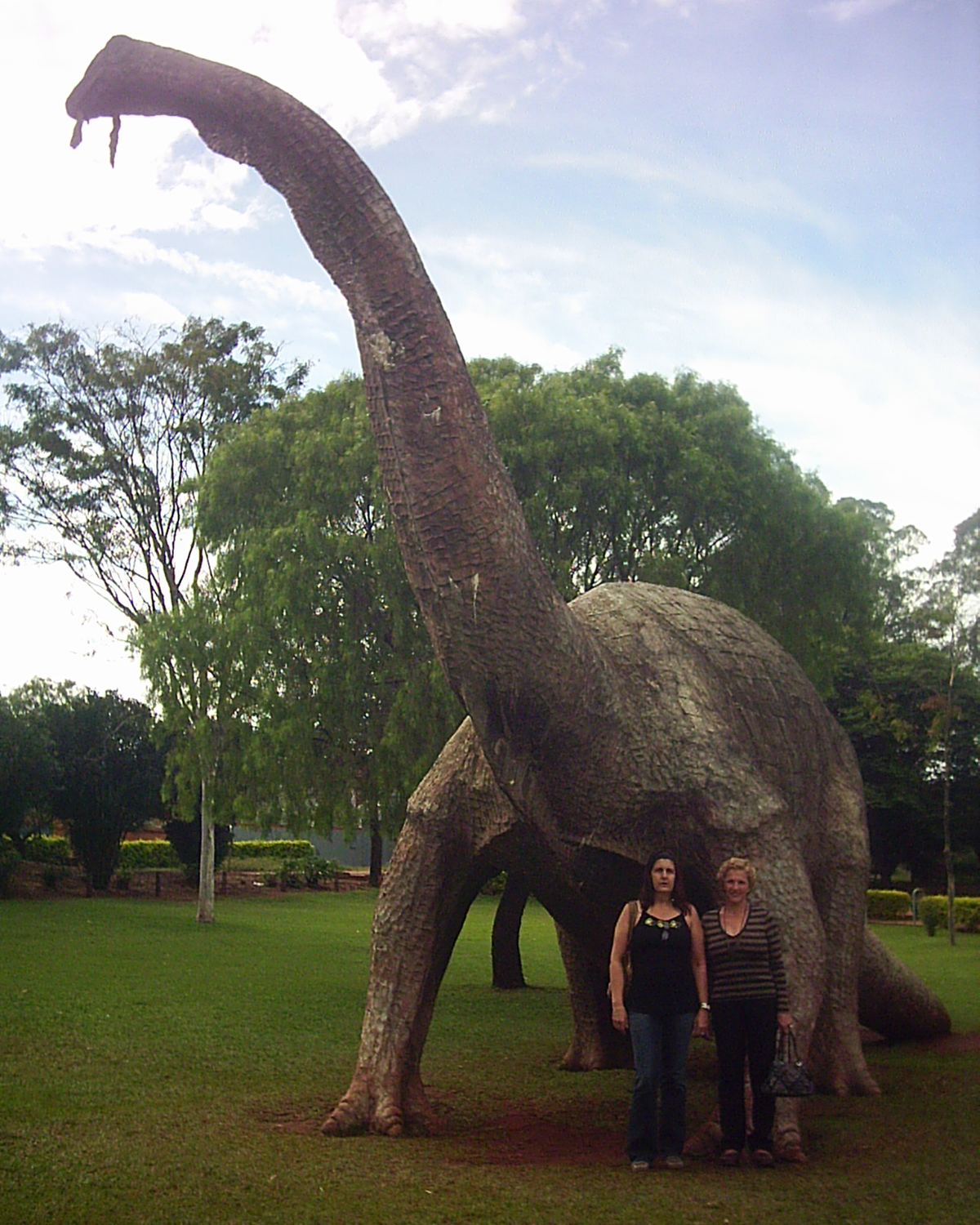 Duas visitantes posando ao pé de uma réplica gigante de dinossauro de pescoço comprido, num gramado arborizado em Peirópolis, Uberaba.