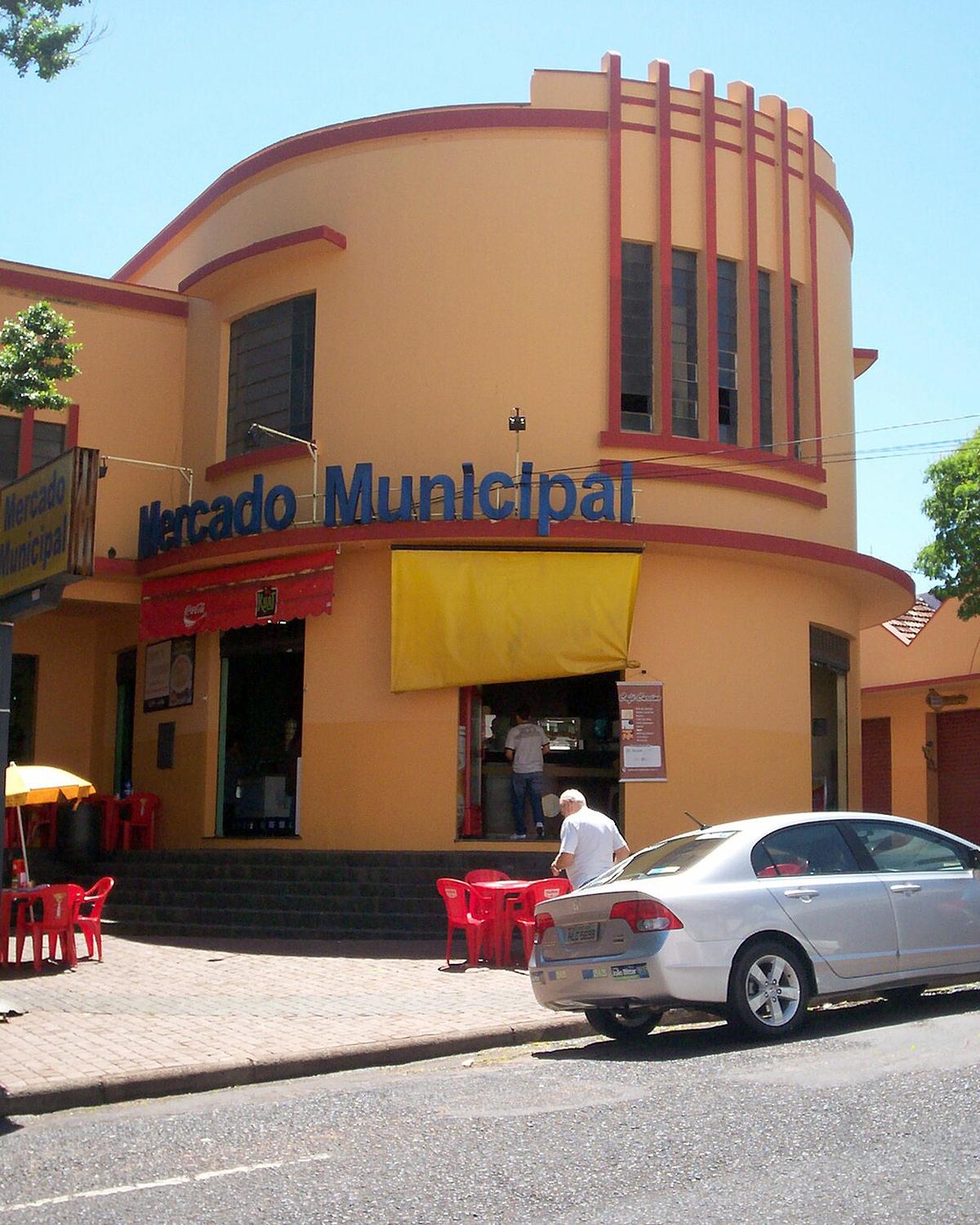 Fachada art déco amarela do Mercado Municipal de Uberlândia, com mesas e cadeiras vermelhas na calçada e um senhor entrando no bar da esquina.