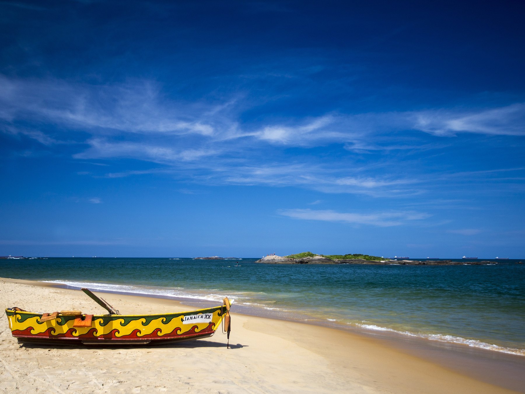 Barco de pesca pintado de amarelo e vermelho parado na areia da Praia de Itapoã, com o mar azul, as pedras da arrebentação e o céu limpo ao fundo.