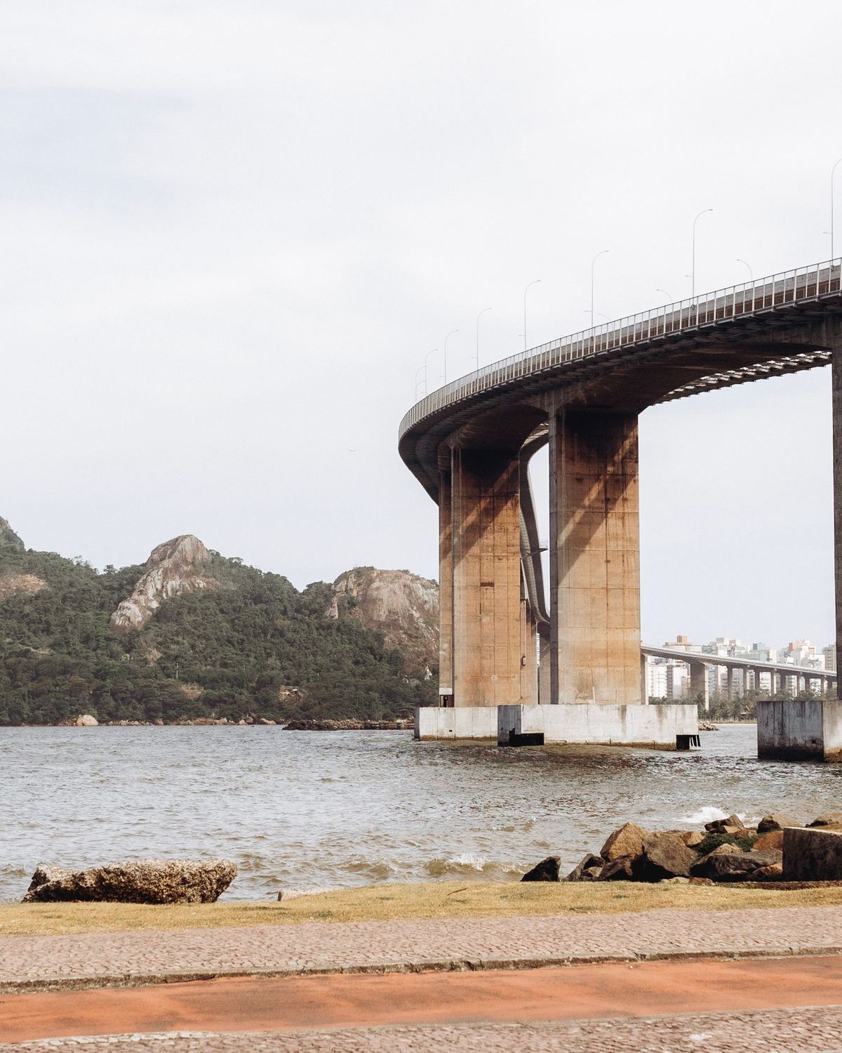 Casal com bicicletas parado na beira da água olhando para a Terceira Ponte, com os pilares da ponte, o morro verde e o calçadão em primeiro plano.