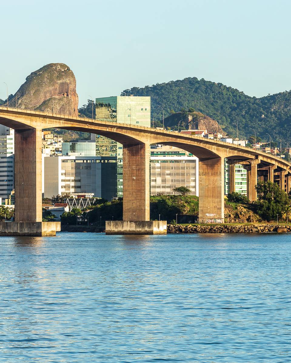 Terceira Ponte vista da água, com os arcos de concreto sobre a baía, os prédios da cidade e um morro de pedra ao fundo em dia de céu azul.
