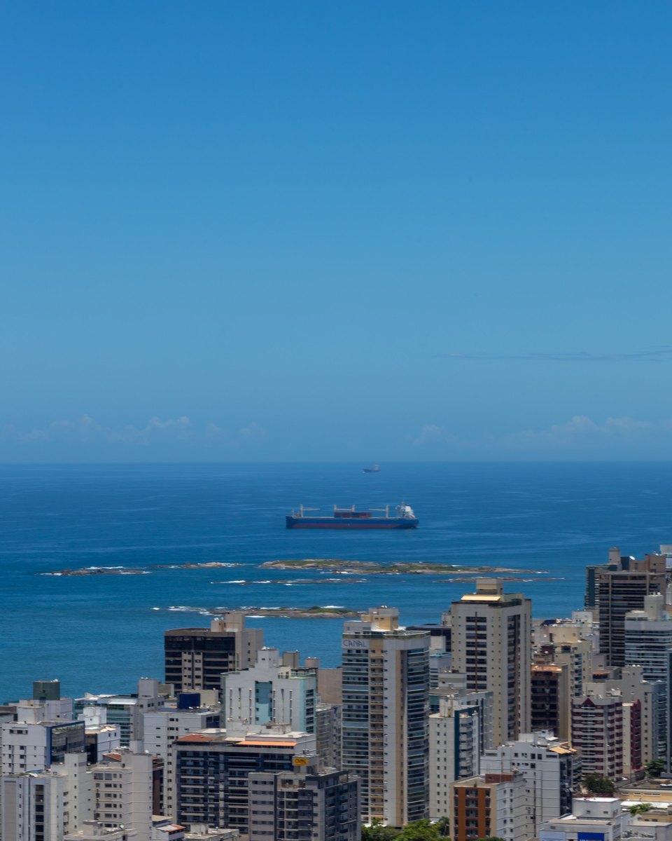Vila Velha vista do alto, com dezenas de prédios altos colados uns nos outros, o mar aberto atrás e um navio cargueiro na linha do horizonte.