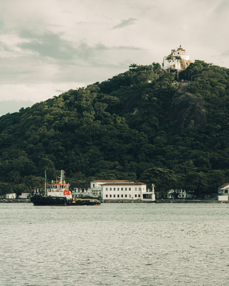 Convento da Penha branco no topo do morro coberto de mata, visto da baía, com casario e uma embarcação atracada na base do morro.