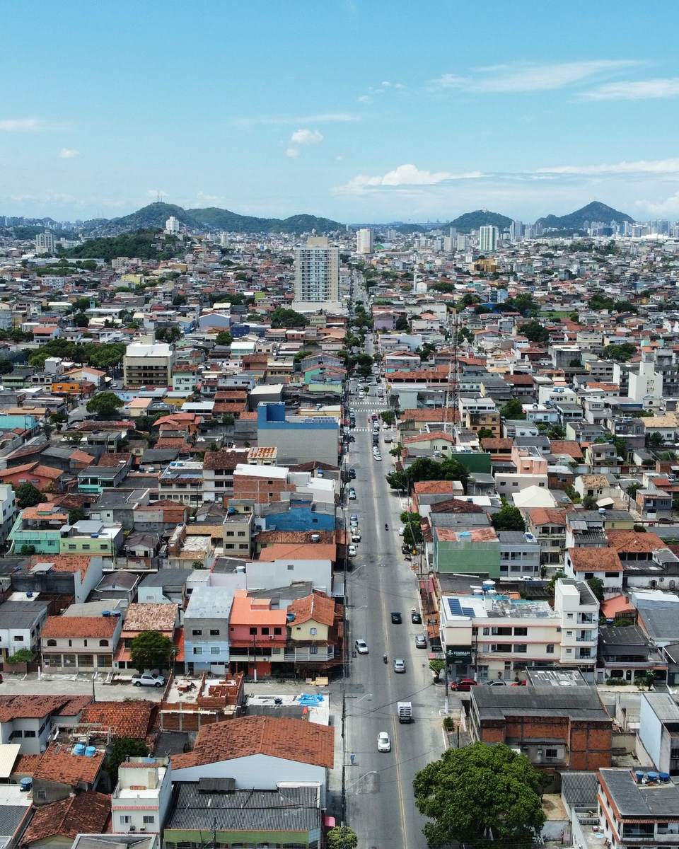 Vista aérea de um bairro residencial de Vila Velha, com telhados vermelhos, uma avenida comercial cortando o meio e os prédios da orla no horizonte.