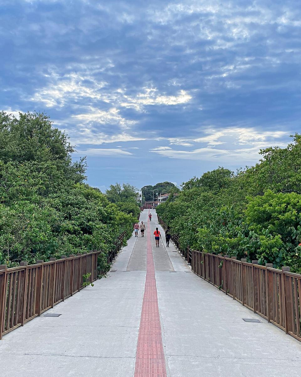 Passarela de concreto com guarda-corpo de madeira atravessando o mangue do Rio Jucu, com pessoas caminhando ao fundo sob céu nublado.