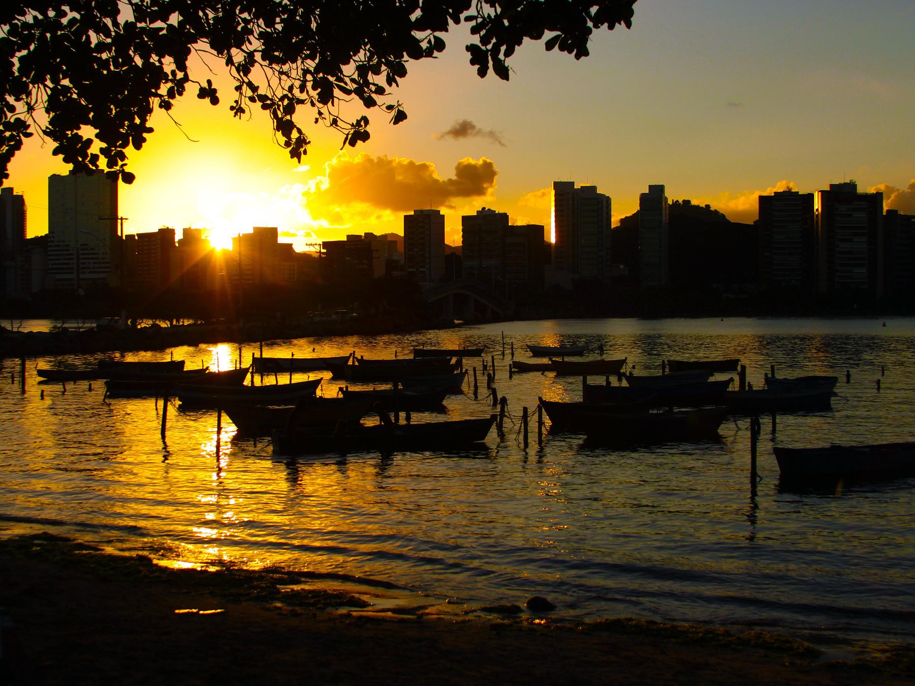 Pôr do sol dourado sobre a baía de Vitória visto da Ilha do Frade, com barcos de pesca ancorados e a silhueta dos prédios da cidade contra o sol.