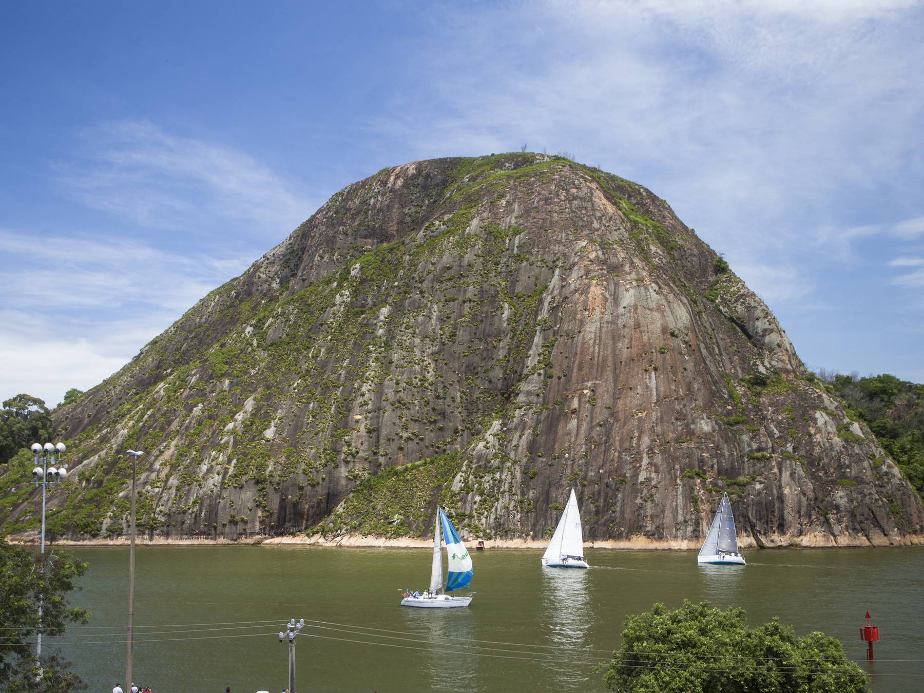 Três veleiros navegando na baía de Vitória em dia de sol, com o paredão de pedra do Morro do Moreno erguido logo atrás.