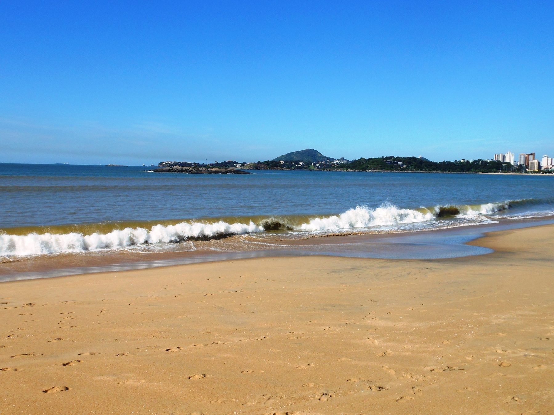 Faixa de areia larga da Praia de Camburi com a onda quebrando, o mar azul e os morros e prédios da cidade na linha do horizonte.